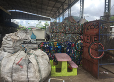 Fotografia contendo diversas sacas de lixo reciclável, à esquerda, uma estante com o lixo organizado, à direita. Em frente a estante, um carrinho de coleta de lixo e uma mesa de plástico com bancos em tamanho infantil.