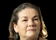 Fotografia com fundo preto mostra uma mulher branca, de cabelos e olhos castanhos.  Ela veste blusa sem mangas na cor verde, usa brincos e colares no mesmo tom.  Ela olha para o lado direito e quase sorri.