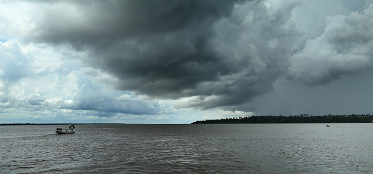 #ParaTodosVerem: Fotografia panorâmica em que se vê um pequeno barco navegando pelo rio. A vegetação ocupa as margens da direita e da esquerda, e o céu está coberto por nuvens de chuva.