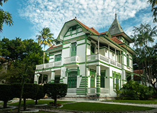 Fotografia de um palacete de dois andares, pintado nas cores verde e branco. Alguns cômodos contam com varanda. O prédio está localizado no centro do terreno e cercado de jardim com espécies nativas da região.