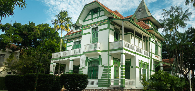 Fotografia de um palacete de dois andares, pintado nas cores verde e branco. Alguns cômodos contam com varanda. O prédio está localizado no centro do terreno e cercado de jardim com espécies nativas da região.