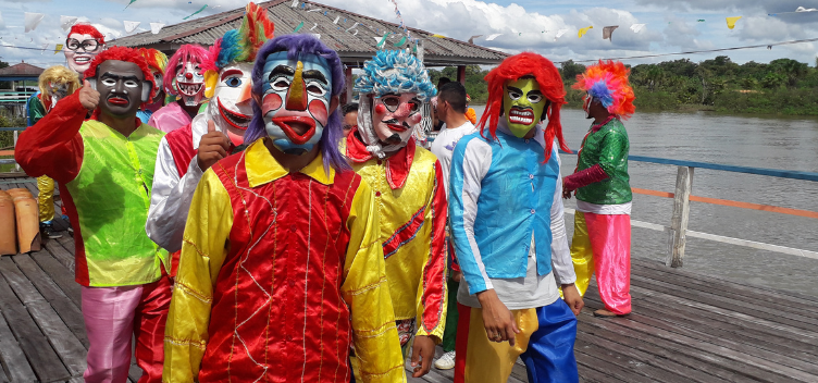 #ParaTodosVerem: Fotografia mostra grupo de pessoas vestindo roupas coloridas e usando máscaras. São brincantes fantasiados para o Carnaval das Águas. Eles estão sobre um trapiche de madeira. Ao fundo, vê-se um rio; e nas margens, a vegetação nativa.