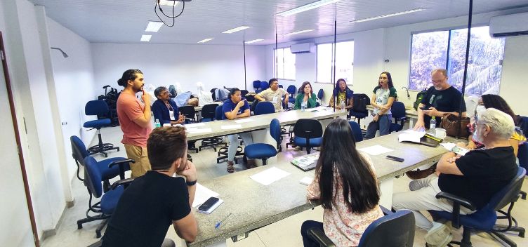 #ParaTodosVerem: Fotografia mostra uma sala de reunião contendo doze pessoas, entre homens e mulheres. Eles estão dispostos em um semicírculo, em que nove pessoas estão sentadas e três em pé. Na sala há dois balcões e diversas cadeiras de cor azul.