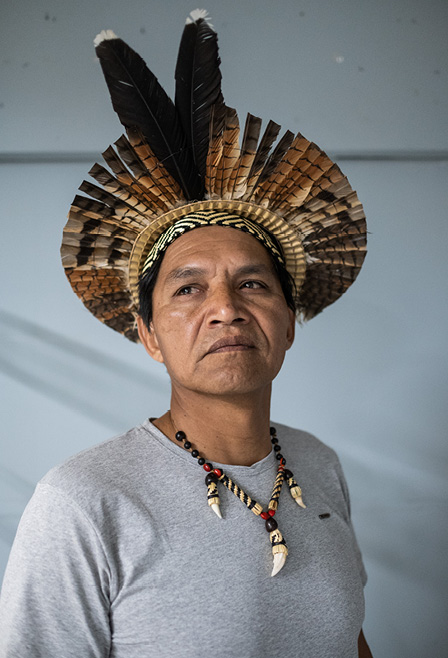 #ParaTodosVerem:  Fotografia com fundo branco mostra um homem indígena. Ele usa cocar e colar, veste uma camiseta cinza de mangas curtas e direciona o olhar para a esquerda.